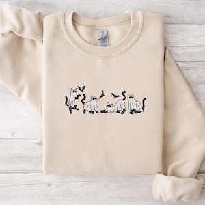 Pode incluir: Camisola bege com gola redonda e mangas compridas. A frente apresenta um desenho bordado de gatos pretos, alguns usando fantasias de fantasmas, com morcegos voando por cima. A camisola est&aacute; dobrada cuidadosamente.