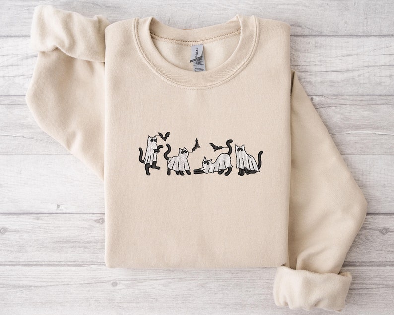 Pode incluir: Camisola bege com gola redonda e mangas compridas. A frente apresenta um desenho bordado de gatos pretos, alguns usando fantasias de fantasmas, com morcegos voando por cima. A camisola est&aacute; dobrada cuidadosamente.
