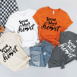 May include: Four t-shirts with the text "Social Work is a work of heart" in black lettering on a white, beige, orange, and gray shirt. The shirts are arranged on a white surface with a pair of blue jeans and a checkered fabric.