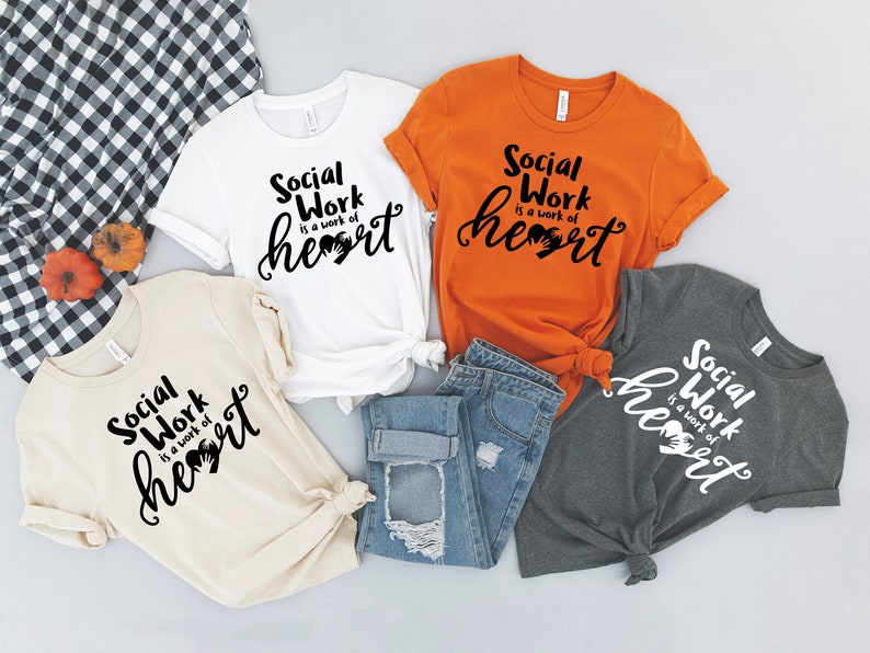 May include: Four t-shirts with the text "Social Work is a work of heart" in black lettering on a white, beige, orange, and gray shirt. The shirts are arranged on a white surface with a pair of blue jeans and a checkered fabric.