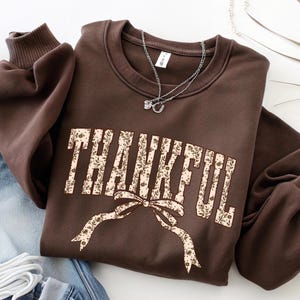 May include: A brown sweatshirt with the word "THANKFUL" in a floral design across the front. A bow with a floral pattern is below the word. A silver necklace is on top of the sweatshirt.