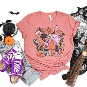 May include: A pink t-shirt with a Halloween-themed design featuring a variety of spooky characters and objects, including a ghost, a witch, a pumpkin, a cauldron, and candy. The text "Trick or Treat" is also included in the design.