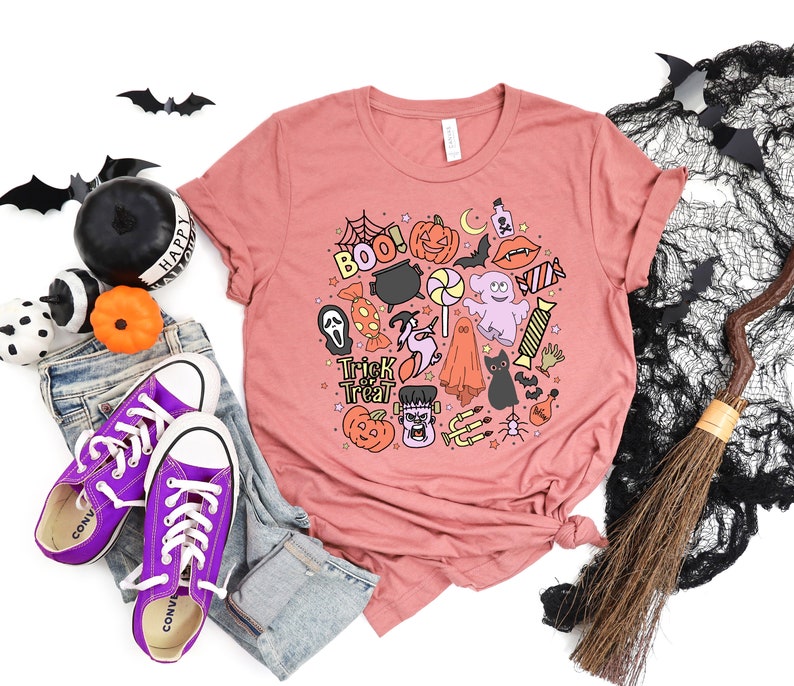 May include: A pink t-shirt with a Halloween-themed design featuring a variety of spooky characters and objects, including a ghost, a witch, a pumpkin, a cauldron, and candy. The text "Trick or Treat" is also included in the design.