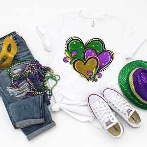 May include: A white t-shirt featuring a design of three hearts in green, purple, and gold with fleur-de-lis accents. The hearts are layered on top of each other. The t-shirt is styled with blue jeans, a gold mask, purple and green beads, white trainers, and a green and purple sequin hat.