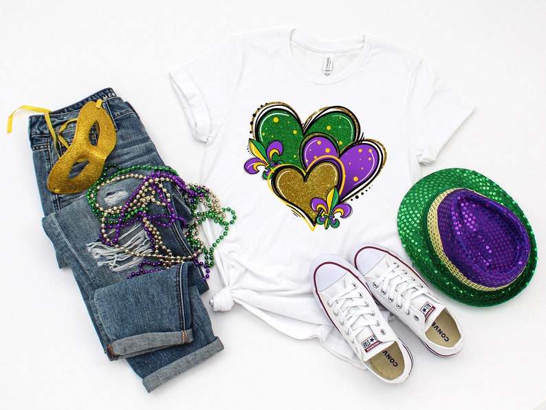 May include: A white t-shirt featuring a design of three hearts in green, purple, and gold with fleur-de-lis accents. The hearts are layered on top of each other. The t-shirt is styled with blue jeans, a gold mask, purple and green beads, white trainers, and a green and purple sequin hat.