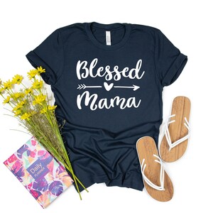 May include: A navy blue t-shirt with the text "Blessed Mama" in white lettering with an arrow and heart graphic. The shirt is laying on a white surface with a floral planner and a pair of white flip flops.