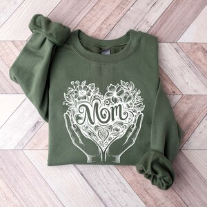 Op de afbeelding: Een groen sweatshirt met een witte afbeelding van een hart met het woord "Mom" erin. Het hart is omringd door bloemen en wordt vastgehouden door twee handen.