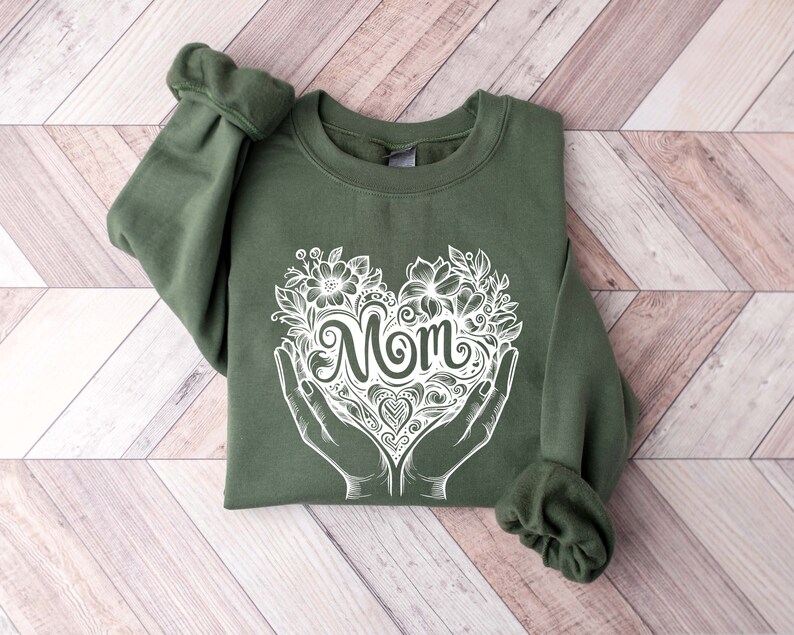 Op de afbeelding: Een groen sweatshirt met een witte afbeelding van een hart met het woord "Mom" erin. Het hart is omringd door bloemen en wordt vastgehouden door twee handen.