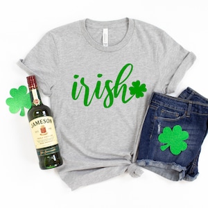May include: A heather gray t-shirt with the word "irish" in green script and a green shamrock. The shirt is paired with denim shorts and a bottle of Jameson Irish Whiskey. A green shamrock is also on the shorts.