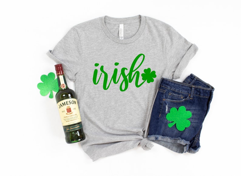 May include: A heather gray t-shirt with the word "irish" in green script and a green shamrock. The shirt is paired with denim shorts and a bottle of Jameson Irish Whiskey. A green shamrock is also on the shorts.