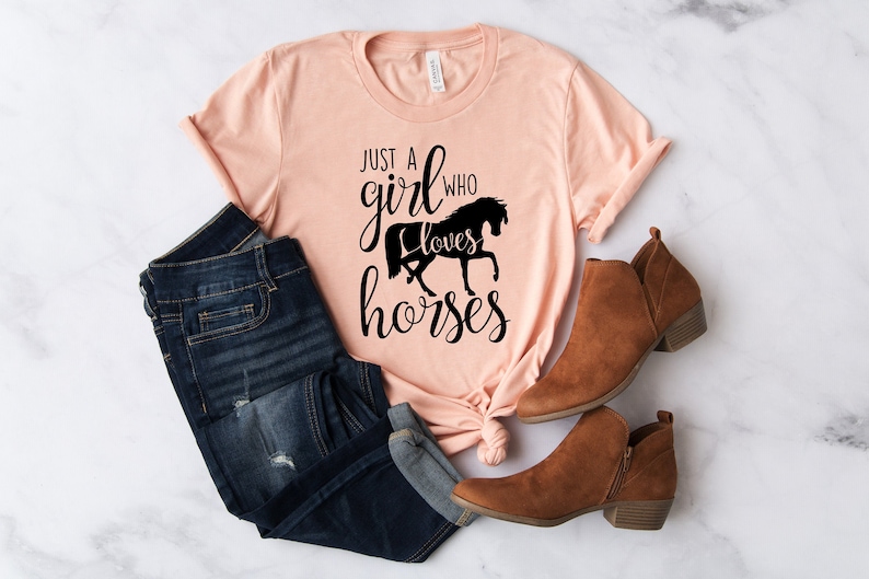 May include: A peach-colored t-shirt with the text "Just a girl who loves horses" in black lettering. The shirt is tied in a knot at the bottom and is paired with blue jeans and brown ankle boots.