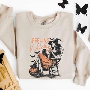 May include: Beige sweatshirt with a graphic of a witch in a hat, sitting in a chair with a black cat and a pumpkin. The text "Feeling Witchy" is above the image. Black butterfly decorations are scattered around the sweatshirt.
