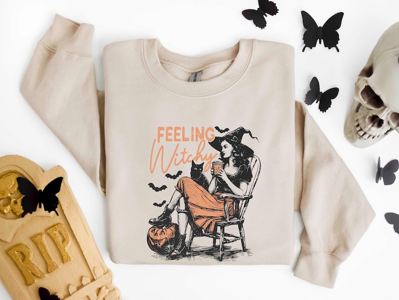 May include: Beige sweatshirt with a graphic of a witch in a hat, sitting in a chair with a black cat and a pumpkin. The text "Feeling Witchy" is above the image. Black butterfly decorations are scattered around the sweatshirt.