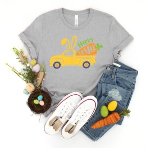 Może przedstawiać: Szary T-shirt z ż&oacute;łtym samochodem dostawczym, kt&oacute;ry przewozi marchewkę z uszami kr&oacute;liczka i napisem "Happy Easter". Koszulka jest połączona z niebieskimi dżinsami, białymi butami sportowymi i gniazdem z jajkami wielkanocnymi.