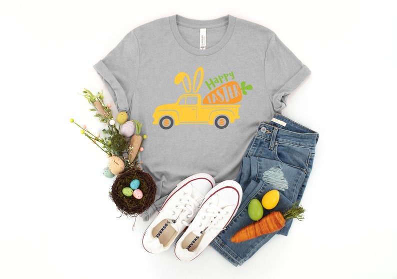Może przedstawiać: Szary T-shirt z ż&oacute;łtym samochodem dostawczym, kt&oacute;ry przewozi marchewkę z uszami kr&oacute;liczka i napisem "Happy Easter". Koszulka jest połączona z niebieskimi dżinsami, białymi butami sportowymi i gniazdem z jajkami wielkanocnymi.