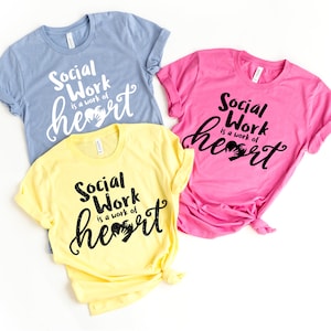 May include: Three t-shirts with the text "Social Work is a work of heart" in black lettering with a handprint heart graphic. The shirts are in blue, yellow, and pink.