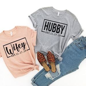May include: Two t-shirts with the words "Wifey" and "Hubby" in black text on a white background. The shirts are on a white surface with a pair of leopard print shoes and blue jeans.