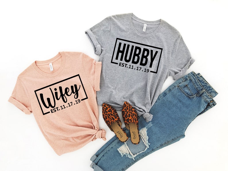May include: Two t-shirts with the words "Wifey" and "Hubby" in black text on a white background. The shirts are on a white surface with a pair of leopard print shoes and blue jeans.