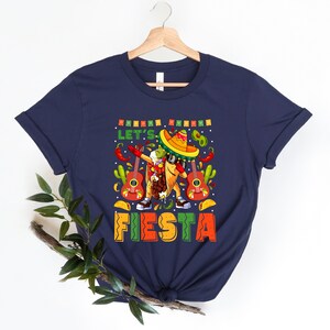 May include: Navy blue t-shirt with a cartoon taco wearing a sombrero and sunglasses. The taco is holding a guitar and the text "Let's Go Fiesta" is printed on the shirt.