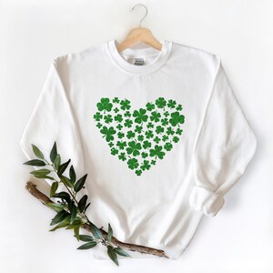 May include: White sweatshirt with a green glitter shamrock heart design. The shamrocks are arranged in a heart shape.