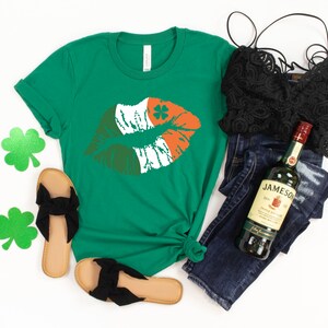 May include: Green t-shirt with a kiss graphic in the colors of the Irish flag, paired with black sandals, a black lace top, and dark wash jeans. A bottle of Jameson Irish Whiskey is also included in the image.