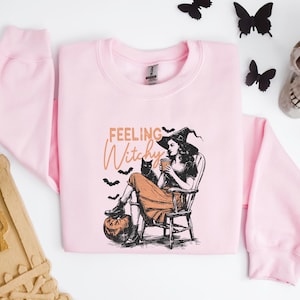 May include: A light pink sweatshirt with a graphic of a witch, a black cat, and the words "Feeling Witchy." The witch is seated, holding a cup, with a pumpkin at her feet. Black bats and butterflies surround the sweatshirt.