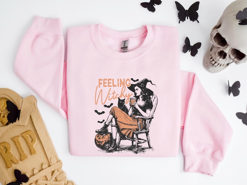 May include: A light pink sweatshirt with a graphic of a witch, a black cat, and the words "Feeling Witchy." The witch is seated, holding a cup, with a pumpkin at her feet. Black bats and butterflies surround the sweatshirt.