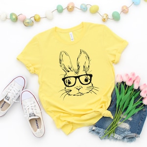 May include: A yellow t-shirt with a black and white illustration of a bunny wearing glasses. The bunny is looking directly at the viewer. The t-shirt is on a white background with a string of pastel Easter eggs and a bouquet of pink tulips.