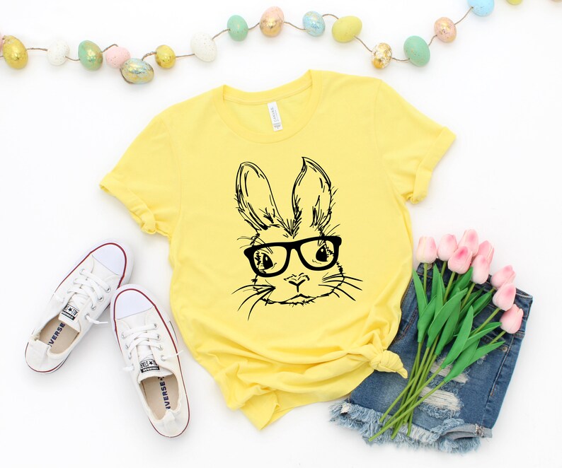 May include: A yellow t-shirt with a black and white illustration of a bunny wearing glasses. The bunny is looking directly at the viewer. The t-shirt is on a white background with a string of pastel Easter eggs and a bouquet of pink tulips.