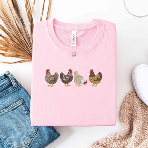 May include: A light pink crew neck t-shirt featuring an embroidered design of four chickens in various colors. The shirt is folded neatly, and a delicate necklace with a small pendant rests on top. A pair of blue jeans and white shoes are in the background.