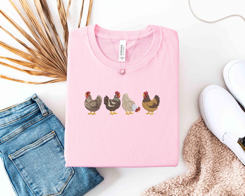 May include: A light pink crew neck t-shirt featuring an embroidered design of four chickens in various colors. The shirt is folded neatly, and a delicate necklace with a small pendant rests on top. A pair of blue jeans and white shoes are in the background.