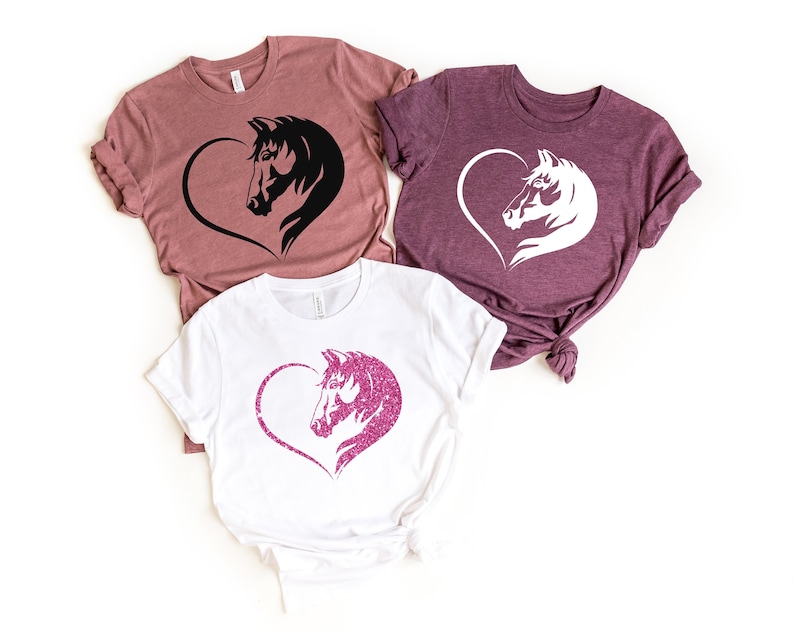 May include: Three t-shirts with a horse head design inside a heart. The shirts are in different colors: mauve, white, and dark purple. The horse head on the white shirt is pink glitter.