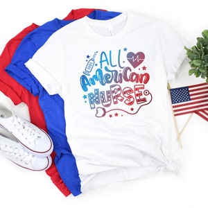 K&ouml;nnte beinhalten: Wei&szlig;es T-Shirt mit dem Text "All American Nurse" in rot, wei&szlig; und blauen Buchstaben. Das Shirt ist gefaltet und wird mit zwei weiteren T-Shirts in rot und blau pr&auml;sentiert.