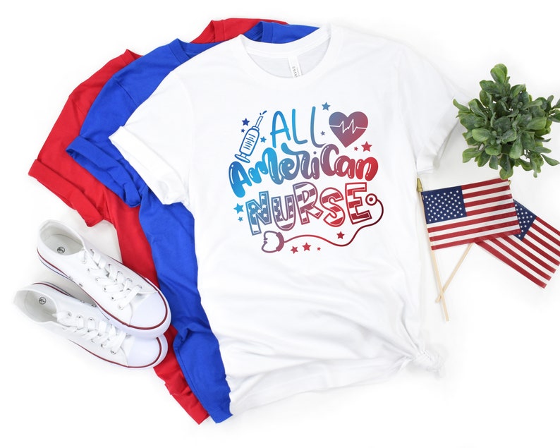 K&ouml;nnte beinhalten: Wei&szlig;es T-Shirt mit dem Text "All American Nurse" in rot, wei&szlig; und blauen Buchstaben. Das Shirt ist gefaltet und wird mit zwei weiteren T-Shirts in rot und blau pr&auml;sentiert.
