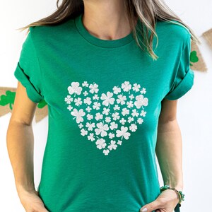 May include: A green t-shirt with a white glitter shamrock heart design. The shirt is a casual style with rolled up sleeves.