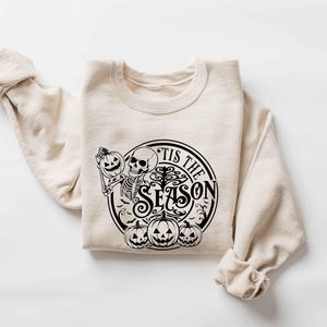 May include: A beige sweatshirt with a black graphic print of a skeleton holding a pumpkin and the text "Tis the Season".