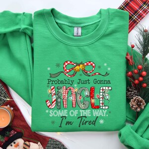 Pu&ograve; includere: Una felpa verde con la scritta "Probably Just Gonna Jingle Some Of The Way. I'm Tired". La parola "JINGLE" &egrave; decorata con luci natalizie e motivi a quadri. Un fiocco a quadri rosso e bianco con campane in cima.