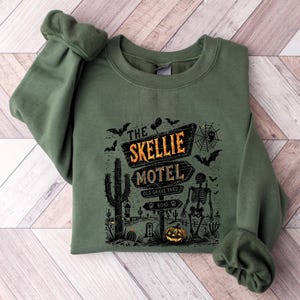 May include: Green sweatshirt featuring a Halloween-themed graphic. The design includes a sign that reads "The Skellie Motel" with a skeleton, bats, a pumpkin, and cacti. The sweatshirt has cuffed sleeves.