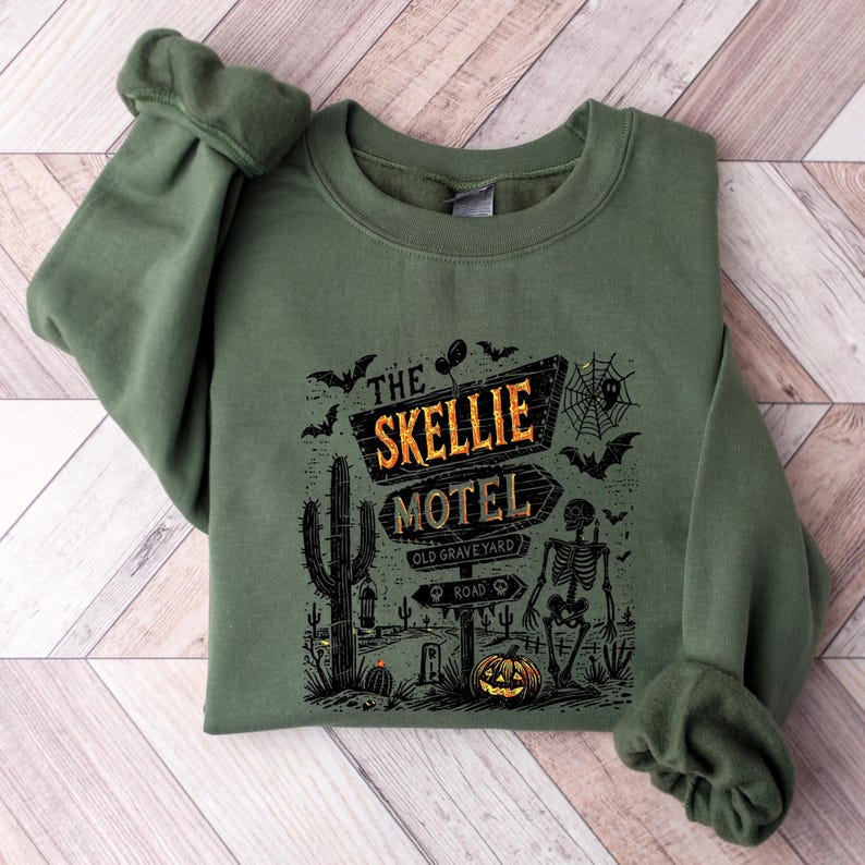 May include: Green sweatshirt featuring a Halloween-themed graphic. The design includes a sign that reads "The Skellie Motel" with a skeleton, bats, a pumpkin, and cacti. The sweatshirt has cuffed sleeves.