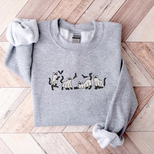 Pode incluir: Um moletom cinza mesclado com um design tem&aacute;tico de Halloween. O design inclui gatos de desenho animado vestidos de fantasmas e morcegos. O moletom &eacute; feito de um material macio e &eacute; adequado para uso casual.