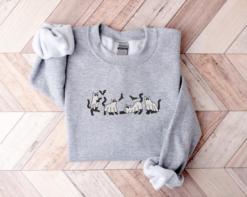 Pode incluir: Um moletom cinza mesclado com um design tem&aacute;tico de Halloween. O design inclui gatos de desenho animado vestidos de fantasmas e morcegos. O moletom &eacute; feito de um material macio e &eacute; adequado para uso casual.