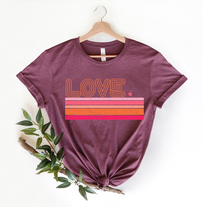 May include: A maroon t-shirt with a retro-style graphic design featuring the word "LOVE" in a bold, outlined font. The graphic also includes a series of horizontal stripes in shades of orange, pink, and red, with a small heart at the end of the stripes.