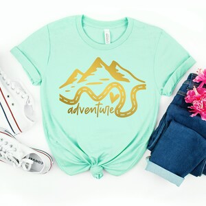 K&ouml;nnte beinhalten: Ein mintgr&uuml;nes T-Shirt mit einer goldenen Grafik von Bergen und einer kurvigen Stra&szlig;e mit einem Herzen. Der Text "adventure" steht unter der Grafik.