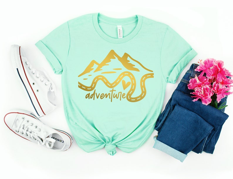 K&ouml;nnte beinhalten: Ein mintgr&uuml;nes T-Shirt mit einer goldenen Grafik von Bergen und einer kurvigen Stra&szlig;e mit einem Herzen. Der Text "adventure" steht unter der Grafik.