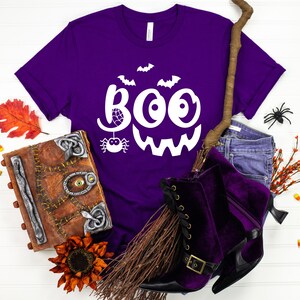 Puede incluir: Camiseta morada con un gr&aacute;fico blanco de "BOO" que presenta una ara&ntilde;a y murci&eacute;lagos. La camiseta se muestra con un sombrero de bruja, una escoba y otras decoraciones de Halloween.
