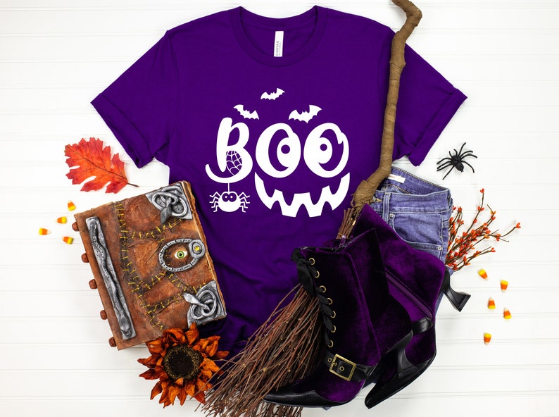 Puede incluir: Camiseta morada con un gr&aacute;fico blanco de "BOO" que presenta una ara&ntilde;a y murci&eacute;lagos. La camiseta se muestra con un sombrero de bruja, una escoba y otras decoraciones de Halloween.
