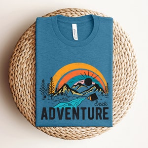 May include: A blue t-shirt with a graphic of a mountain landscape, rainbow, and the words "Seek Adventure." The design includes a river, trees, and a tent. The shirt is folded on a woven surface.