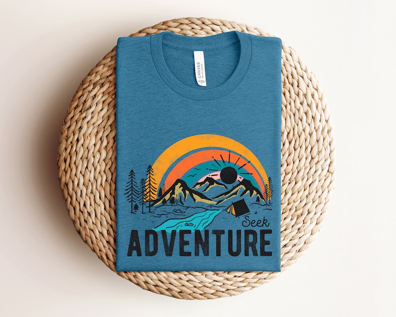 May include: A blue t-shirt with a graphic of a mountain landscape, rainbow, and the words "Seek Adventure." The design includes a river, trees, and a tent. The shirt is folded on a woven surface.