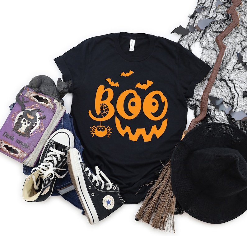 Puede incluir: Camiseta negra con un dise&ntilde;o de cara de calabaza naranja y la palabra "BOO" en una fuente estilizada. El dise&ntilde;o incluye una ara&ntilde;a y murci&eacute;lagos.