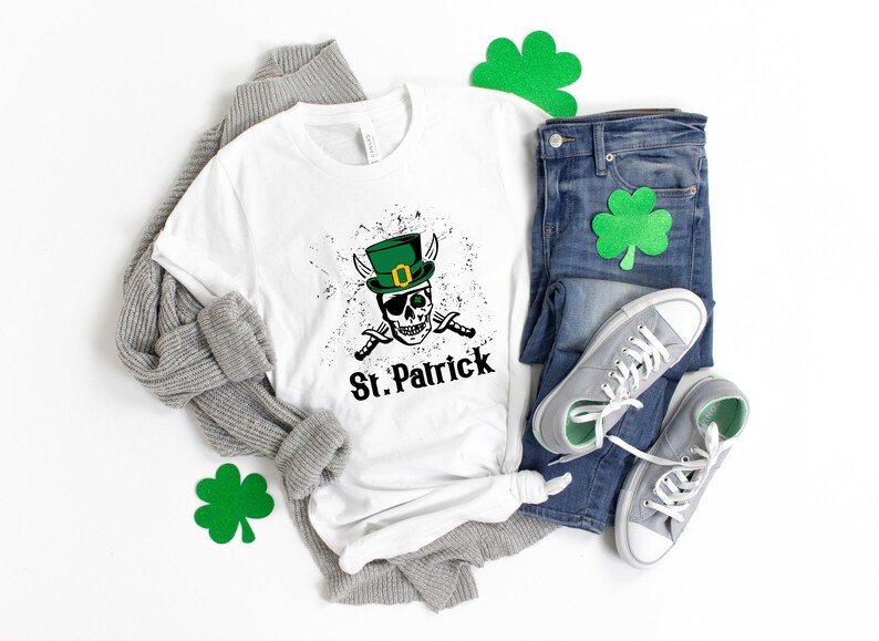 May include: White t-shirt with a green leprechaun skull graphic and the text "St. Patrick". The shirt is laid out on a white surface with a gray sweater, blue jeans, and green shamrock decorations.